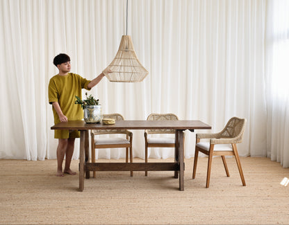 Person adjusting a pendant light over a wooden dining table with chairs in a room with white curtains.