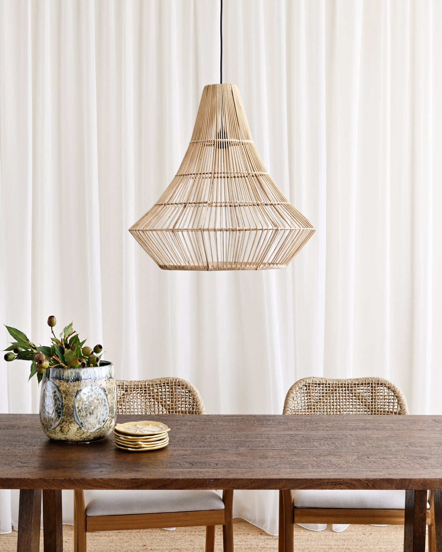 Dining area with a wooden table, chairs, and a wicker pendant light against a white curtain background.