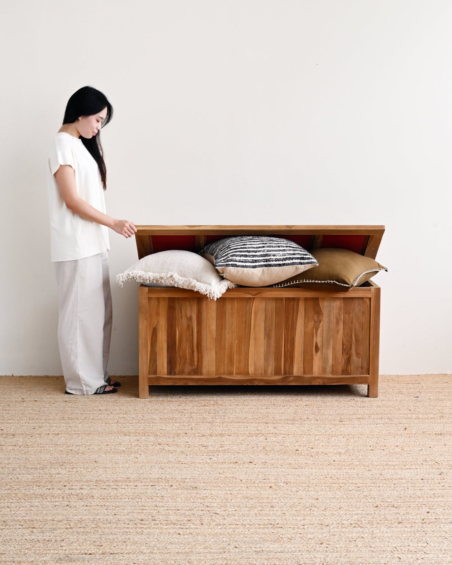Woman opening a wooden storage chest with pillows inside on a plain floor.