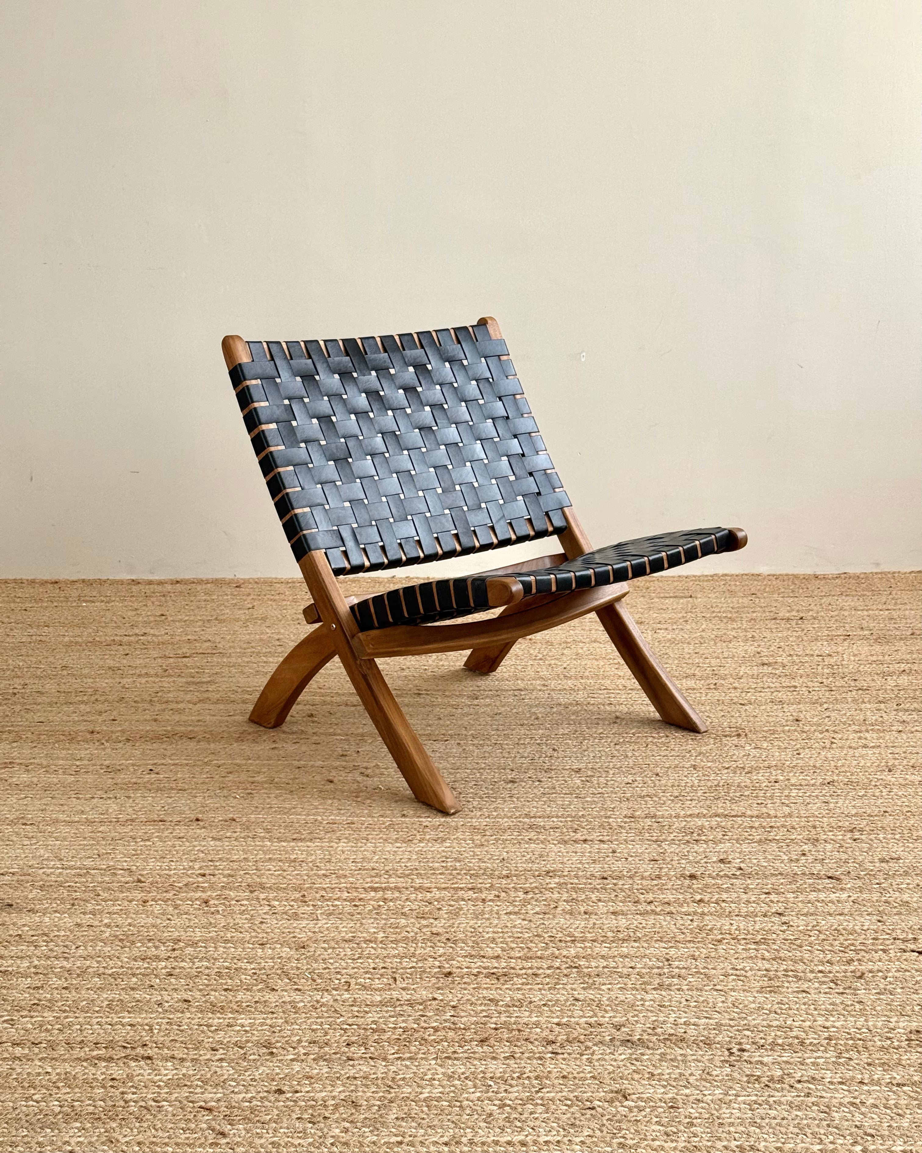 A mid-century modern wooden lounge chair with woven blue-gray leather straps, set on a textured beige jute rug against a plain beige wall.