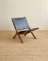 A mid-century modern wooden lounge chair with woven blue-gray leather straps, set on a textured beige jute rug against a plain beige wall.