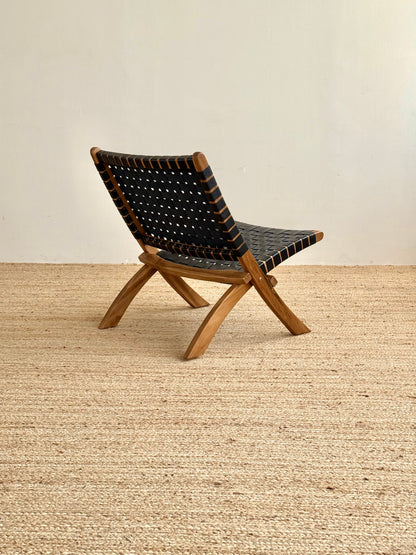A mid-century modern folding chair with a natural wood frame and black woven leather straps, shown on a textured jute rug against a neutral beige wall.