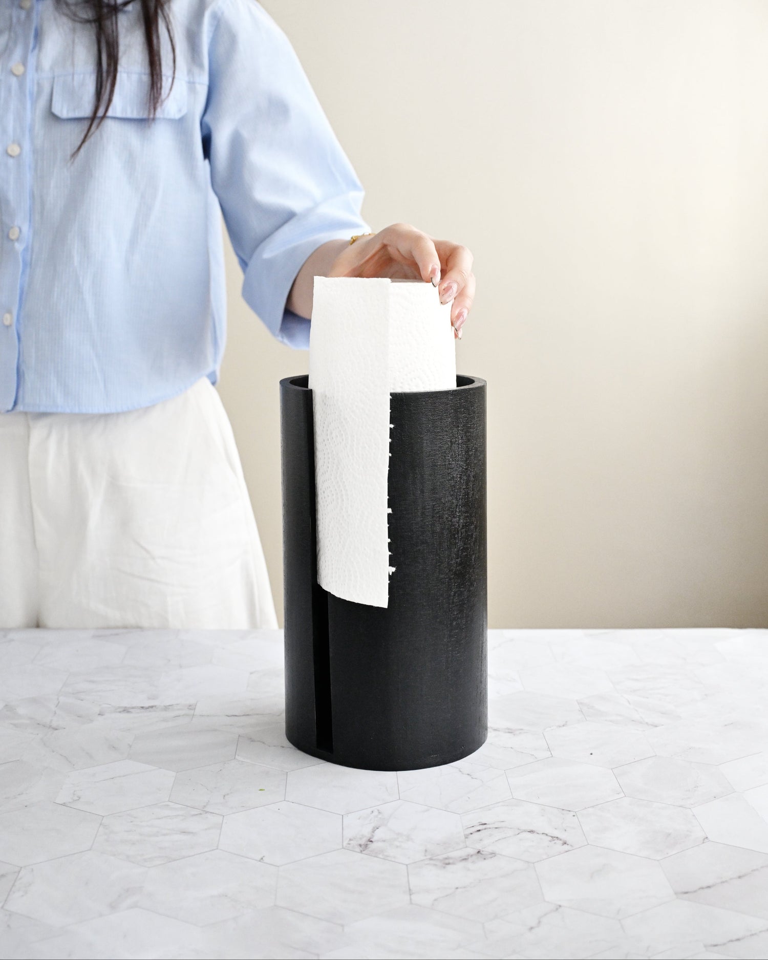 Person holding a white paper towel above a black trash can on a light-colored surface.