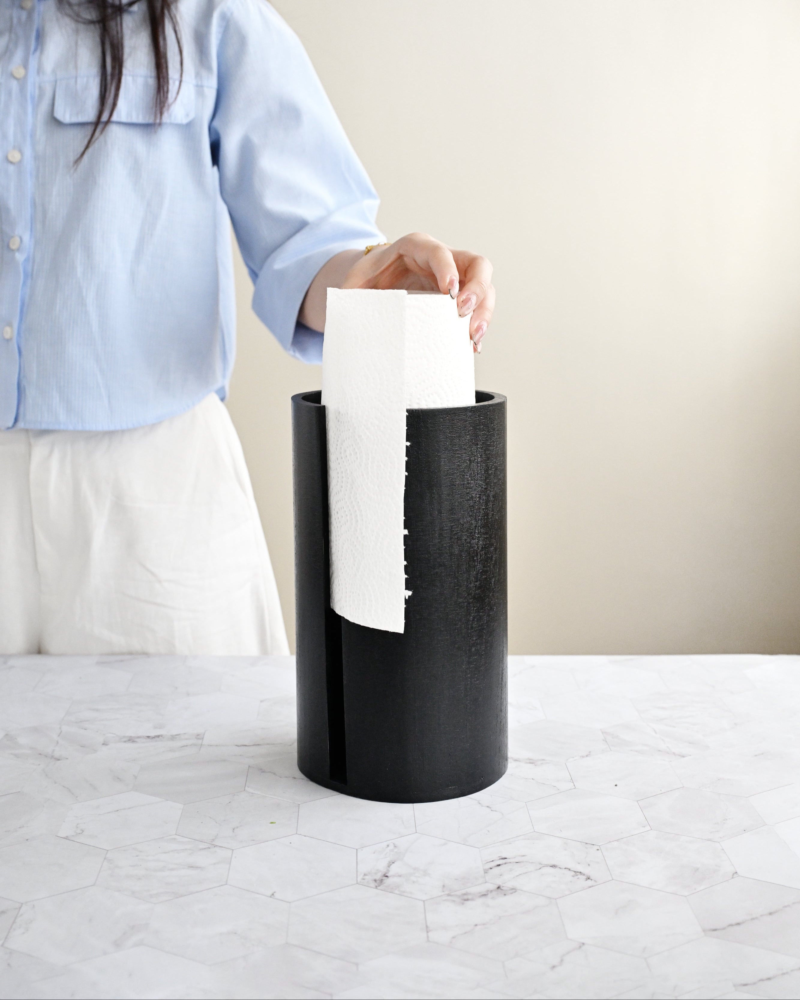 Person holding a white paper towel above a black trash can on a light-colored surface.