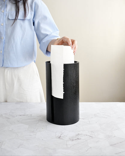 Person holding a white paper towel above a black trash can on a light-colored surface.