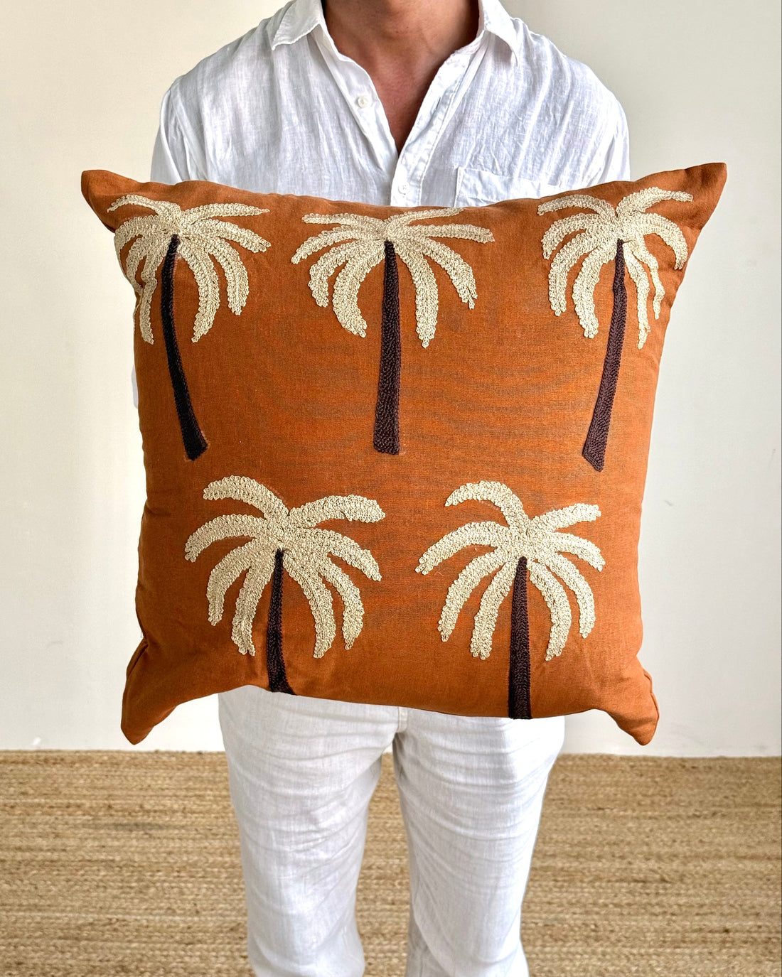 A person wearing a white shirt and white pants holds a square throw pillow with a rust-orange background and cream and dark brown palm tree motifs.