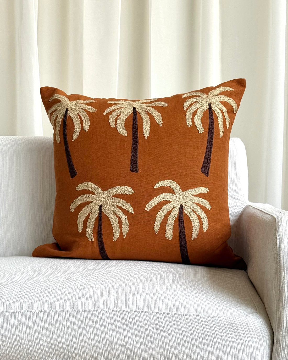 An orange-brown throw pillow with a palm tree pattern, resting on a light gray textured sofa in front of cream curtains.