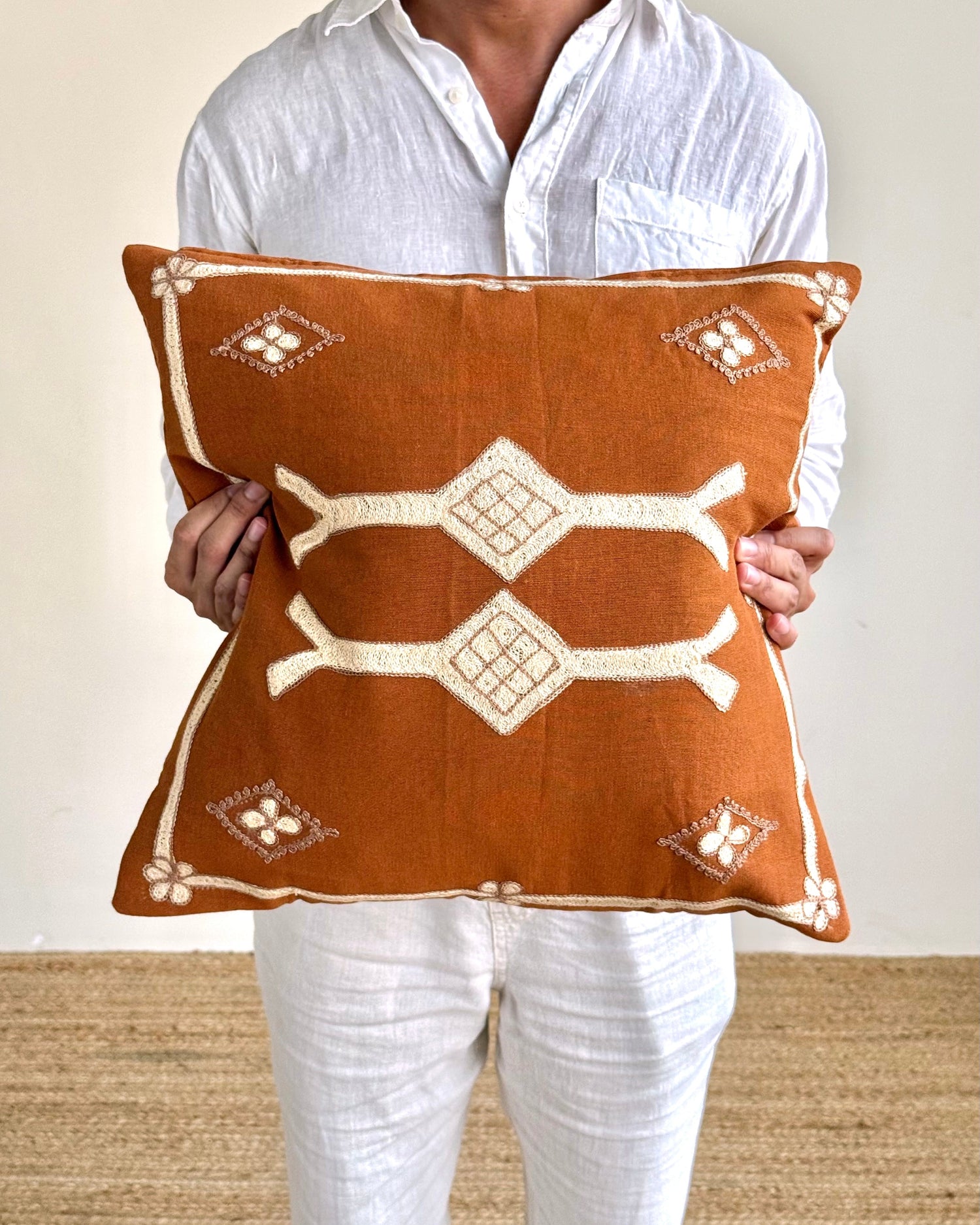 A person in a white shirt and white pants holds a square brown throw pillow with cream-colored geometric patterns and borders.