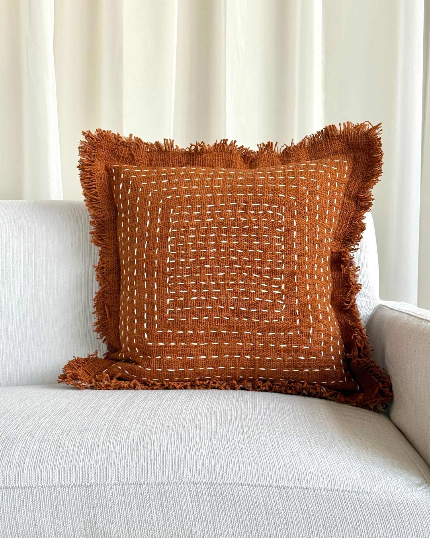 A square brown woven throw pillow with fringed edges and white stitched geometric patterns, resting on a light gray textured sofa in front of white curtains.