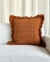 A square brown woven throw pillow with fringed edges and white stitched geometric patterns, resting on a light gray textured sofa in front of white curtains.