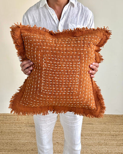 A person wearing a white shirt and white pants holds a square rust-brown woven throw pillow with fringed edges against a plain beige wall and light woven rug.
