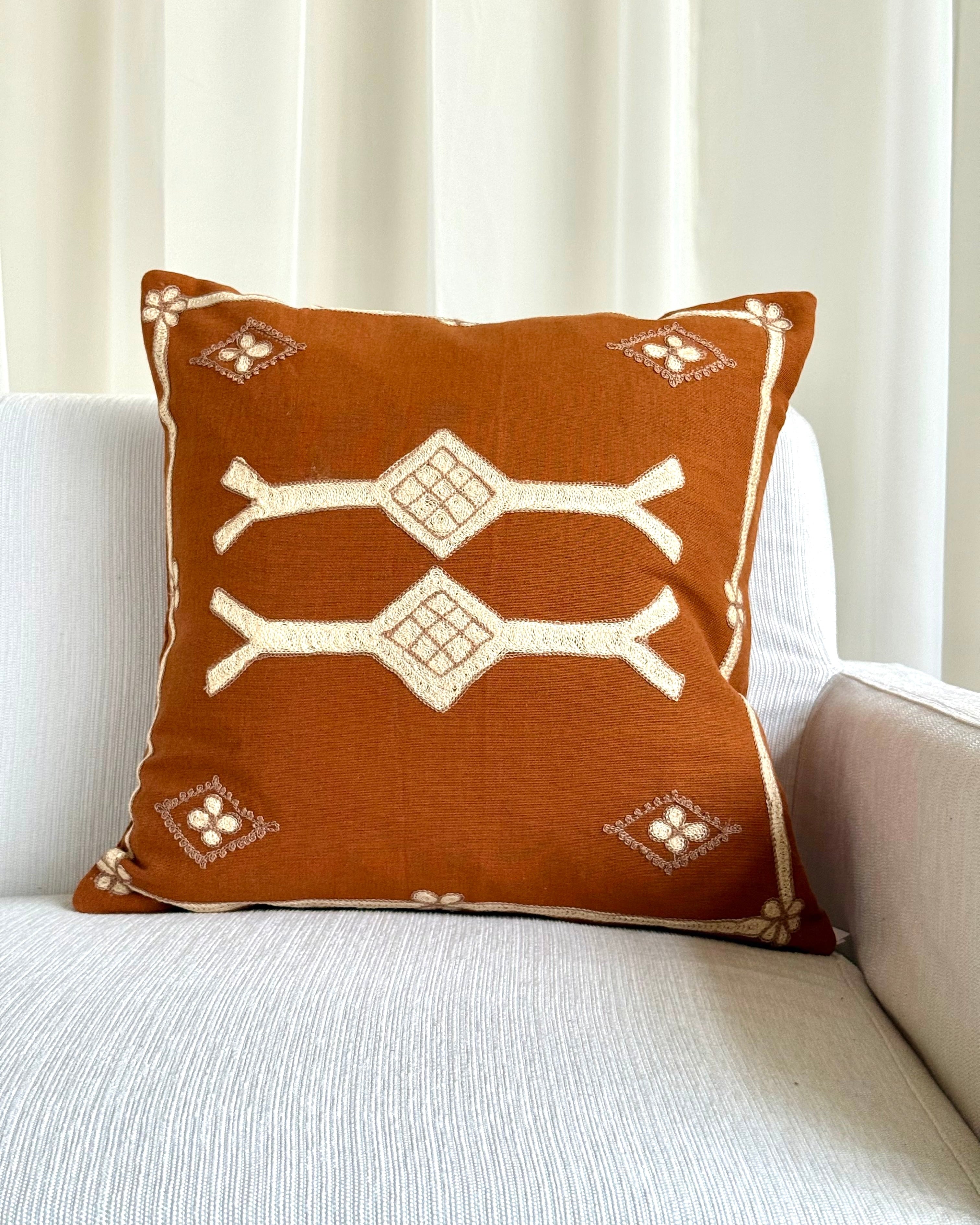 A square throw pillow in rust brown with cream-colored geometric and tribal-style patterns, resting on a light gray textured sofa in front of a white curtain.