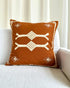 A square throw pillow in rust brown with cream-colored geometric and tribal-style patterns, resting on a light gray textured sofa in front of a white curtain.