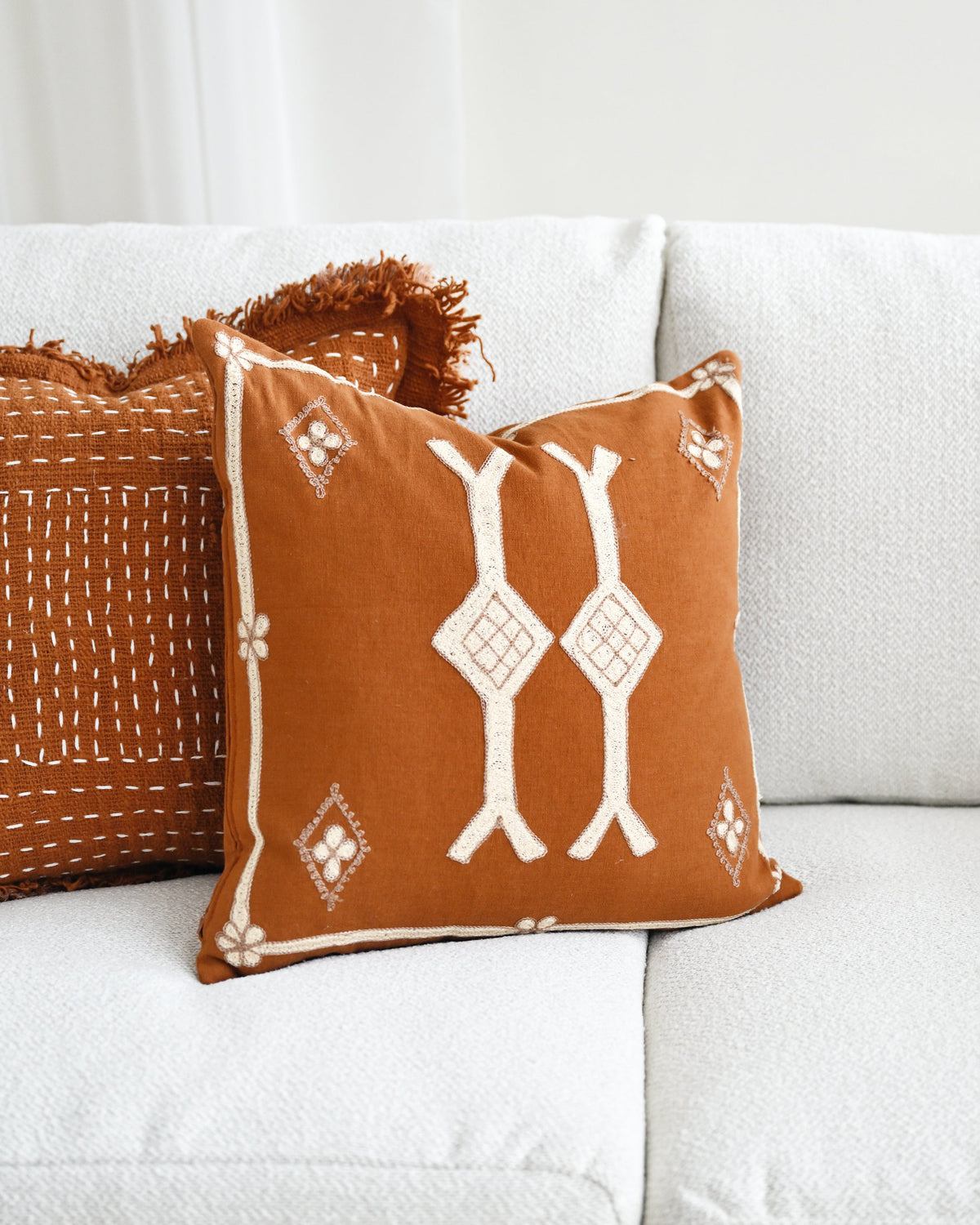 A rust-orange throw pillow with white geometric and tribal-style patterns sits on a white textured sofa next to another orange pillow with white stitching.