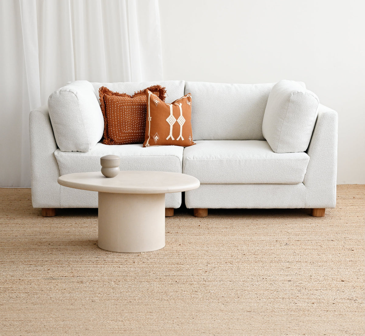 A modern white sectional sofa in a bright, minimalist living room setting, styled with two textured accent pillows in rust orange and cream, a round low coffee table, and a light beige jute rug.