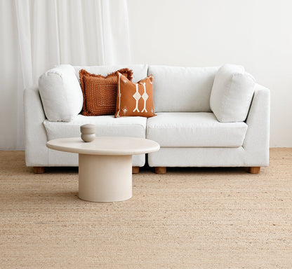 A modern white sectional sofa in a bright, minimalist living room setting, styled with two textured accent pillows in rust orange and cream, a round low coffee table, and a light beige jute rug.