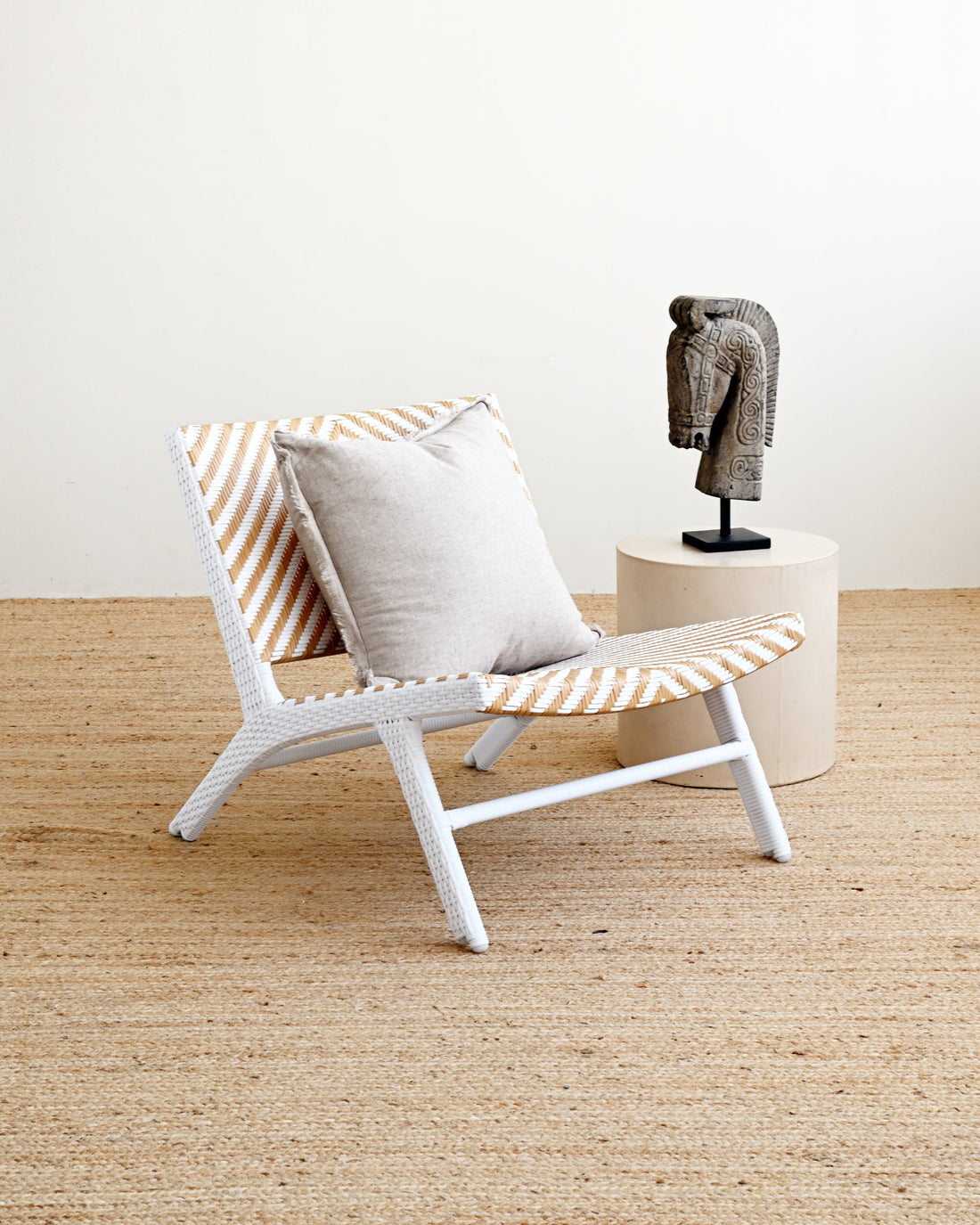 A modern white woven armchair with orange and white striped cushions, styled with a light gray throw pillow on a natural jute rug. A small beige side table with a gray sculptural bust is positioned behind the chair against a clean off-white wall.