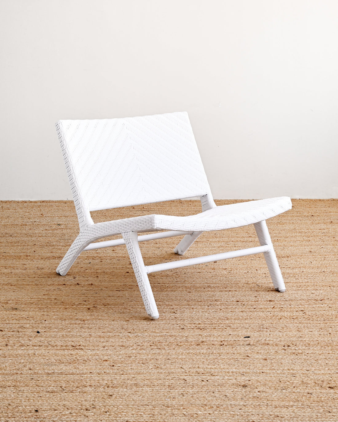 A white woven outdoor lounge chair with a curved backrest and seat, positioned on a natural jute rug against a plain off-white wall.