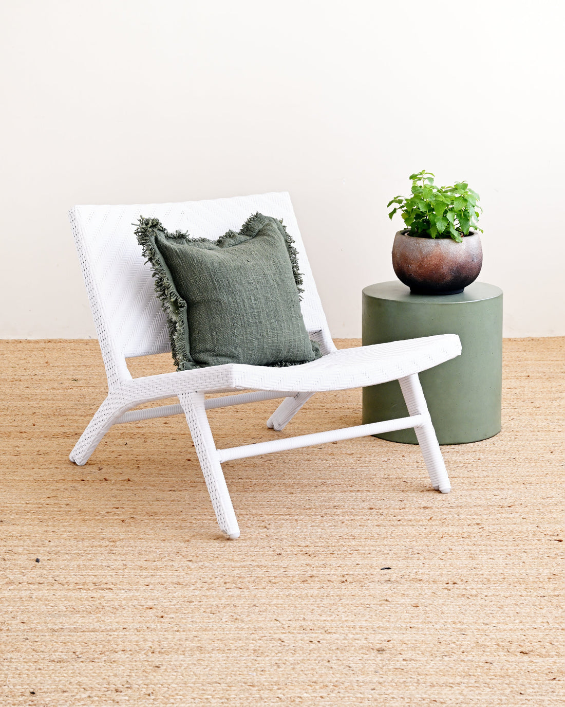 A white woven outdoor lounge chair with a dark green textured throw pillow, set on a natural jute rug next to a green cylindrical side table holding a potted plant.