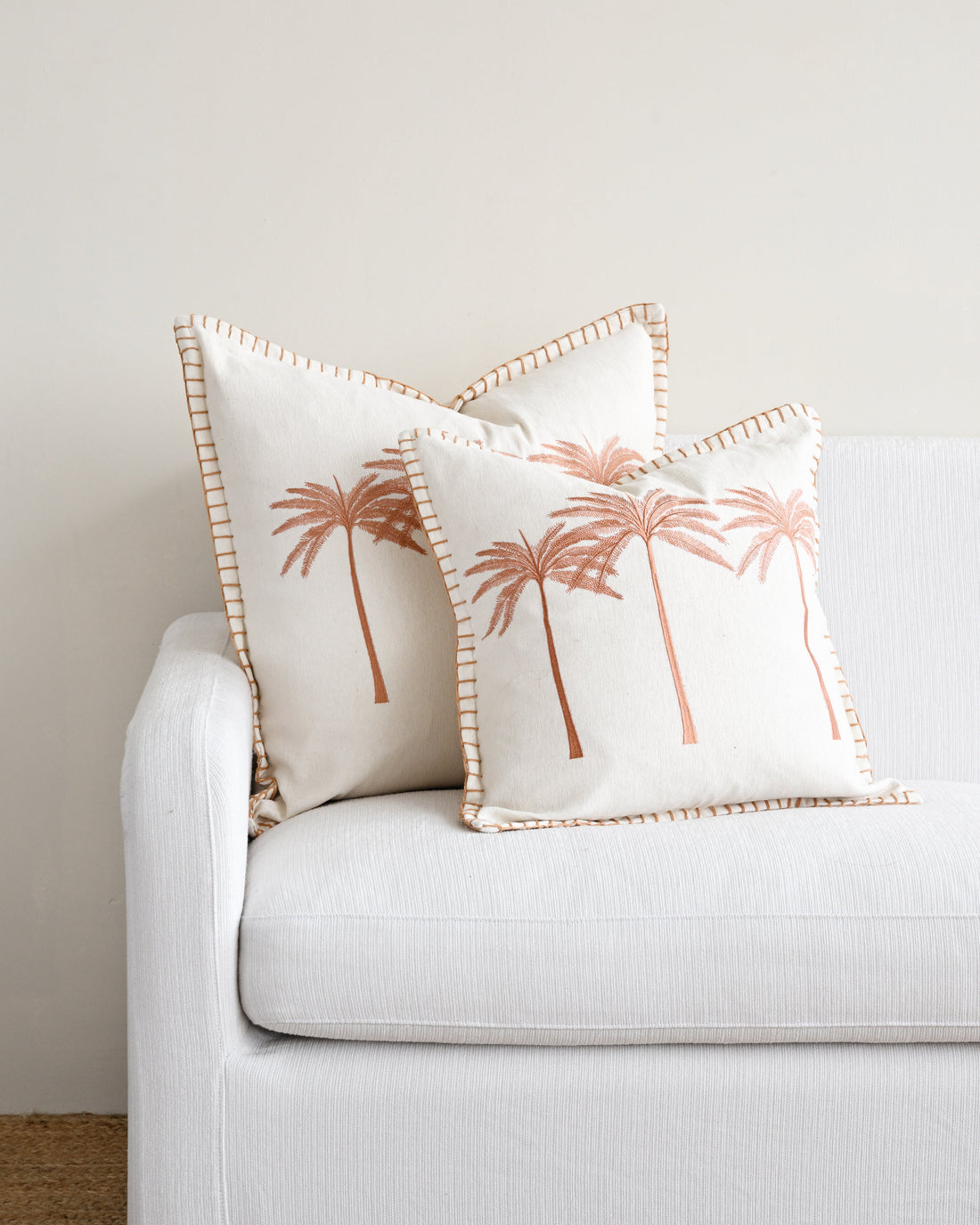 Two decorative pillows with palm tree designs on a white couch against a light gray wall.