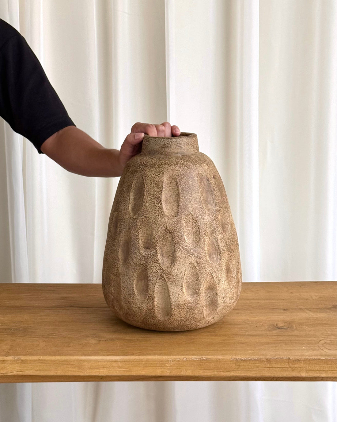 A textured, organic-shaped ceramic vase in a sandy beige color sits on a wooden table, with a person&
