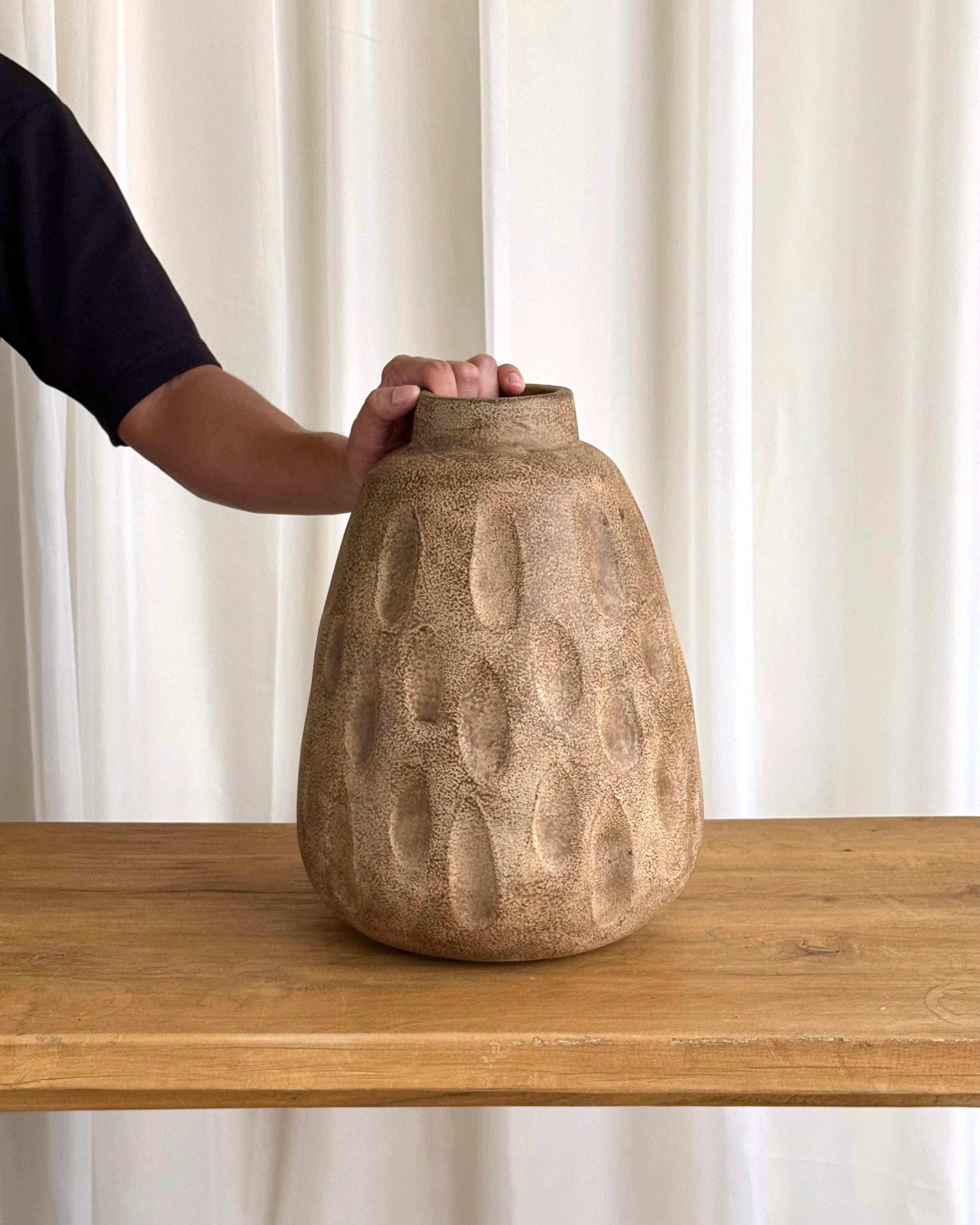 A textured, organic-shaped ceramic vase in a sandy beige color sits on a wooden table, with a person&