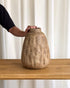 A textured, organic-shaped ceramic vase in a sandy beige color sits on a wooden table, with a person&