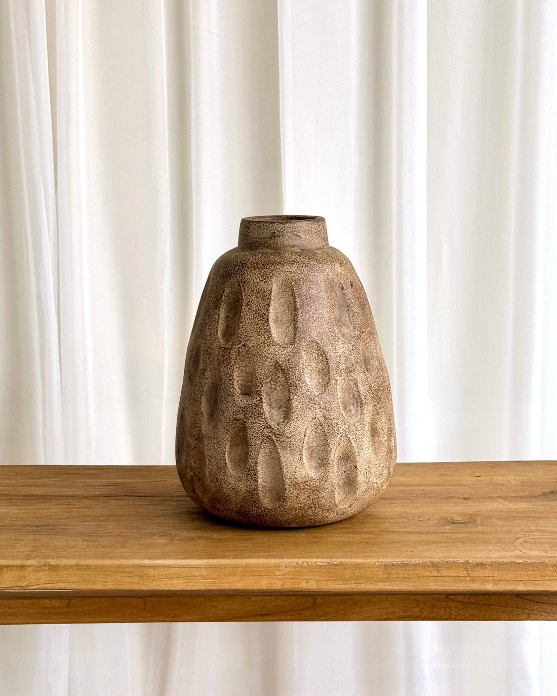 A textured, organic-shaped ceramic vase in a warm brown and beige tone, sitting on a light wood table against a white curtain background.