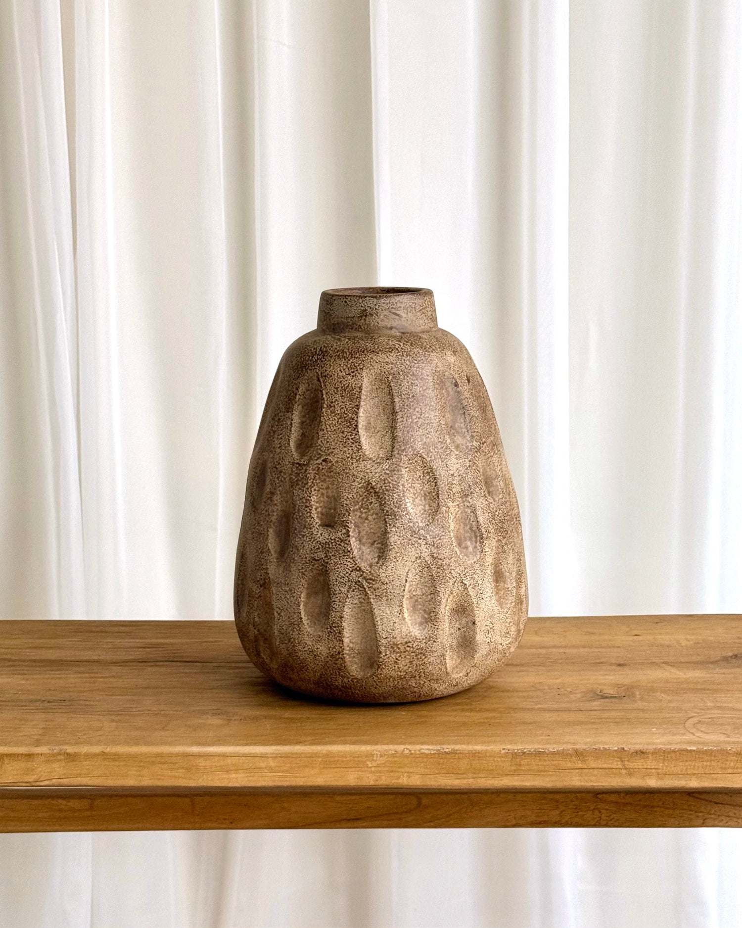 A textured, organic-shaped ceramic vase in a warm brown and beige tone, sitting on a light wood table against a white curtain background.