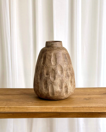 A textured, organic-shaped ceramic vase in a warm brown and beige tone, sitting on a light wood table against a white curtain background.