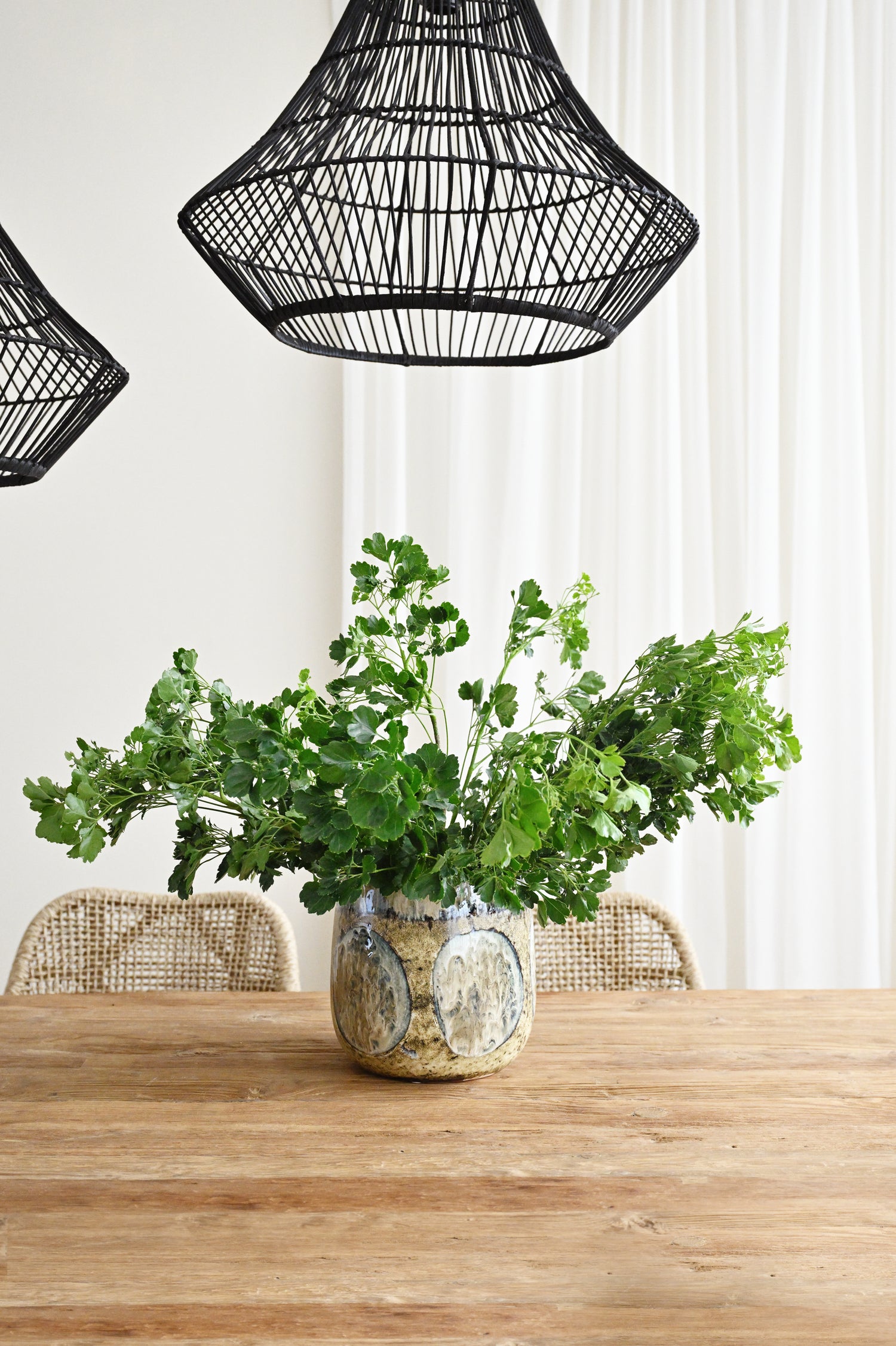 Vase with green plants on a wooden table under black wicker pendant lights.