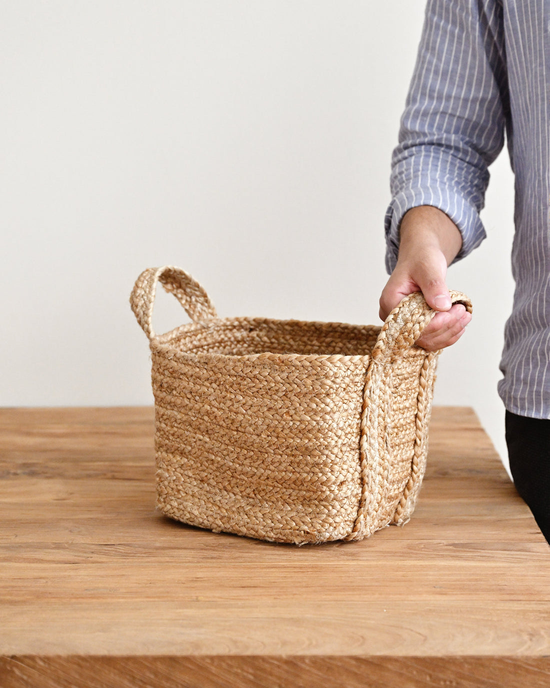 A woven natural fiber storage basket with two handles sits on a wooden table, with a person&
