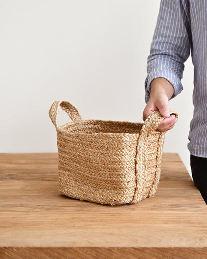 A woven natural fiber storage basket with two handles sits on a wooden table, with a person&