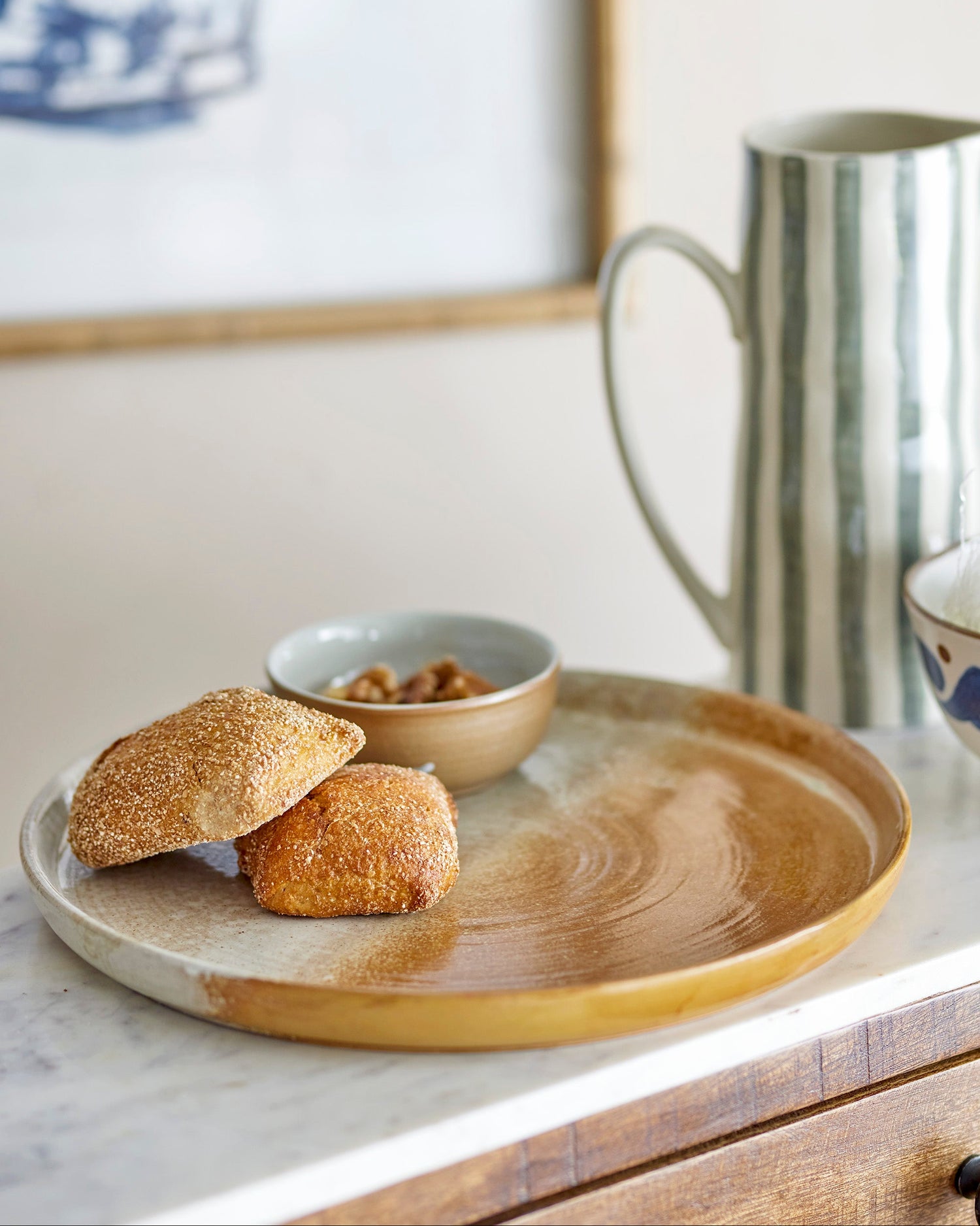 Two bread rolls on a wooden plate with a bowl of nuts and a metallic pitcher in the background.