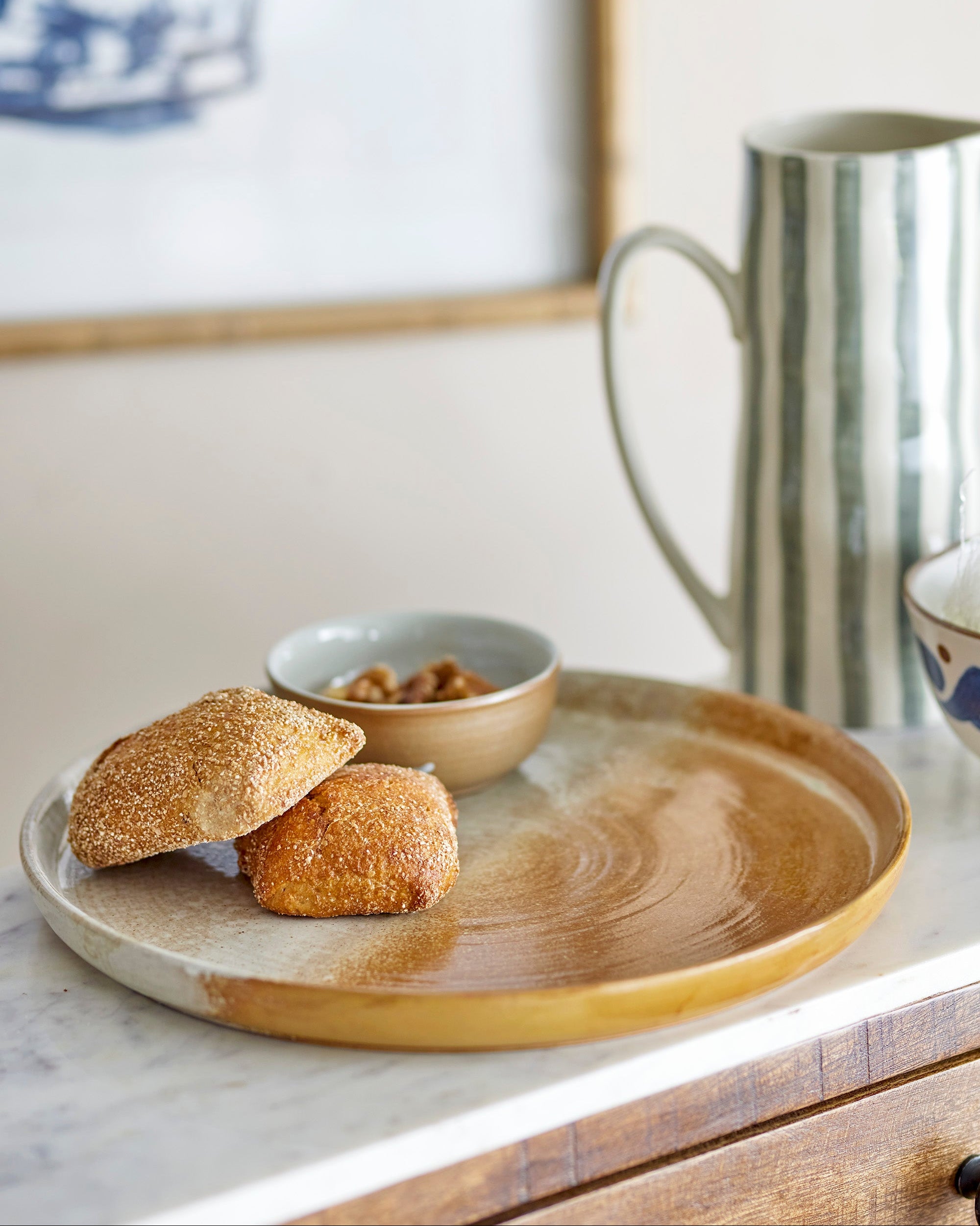 Two bread rolls on a wooden plate with a bowl of nuts and a metallic pitcher in the background.