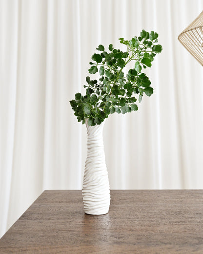 White textured vase with greenery on a wooden surface against a white curtain background