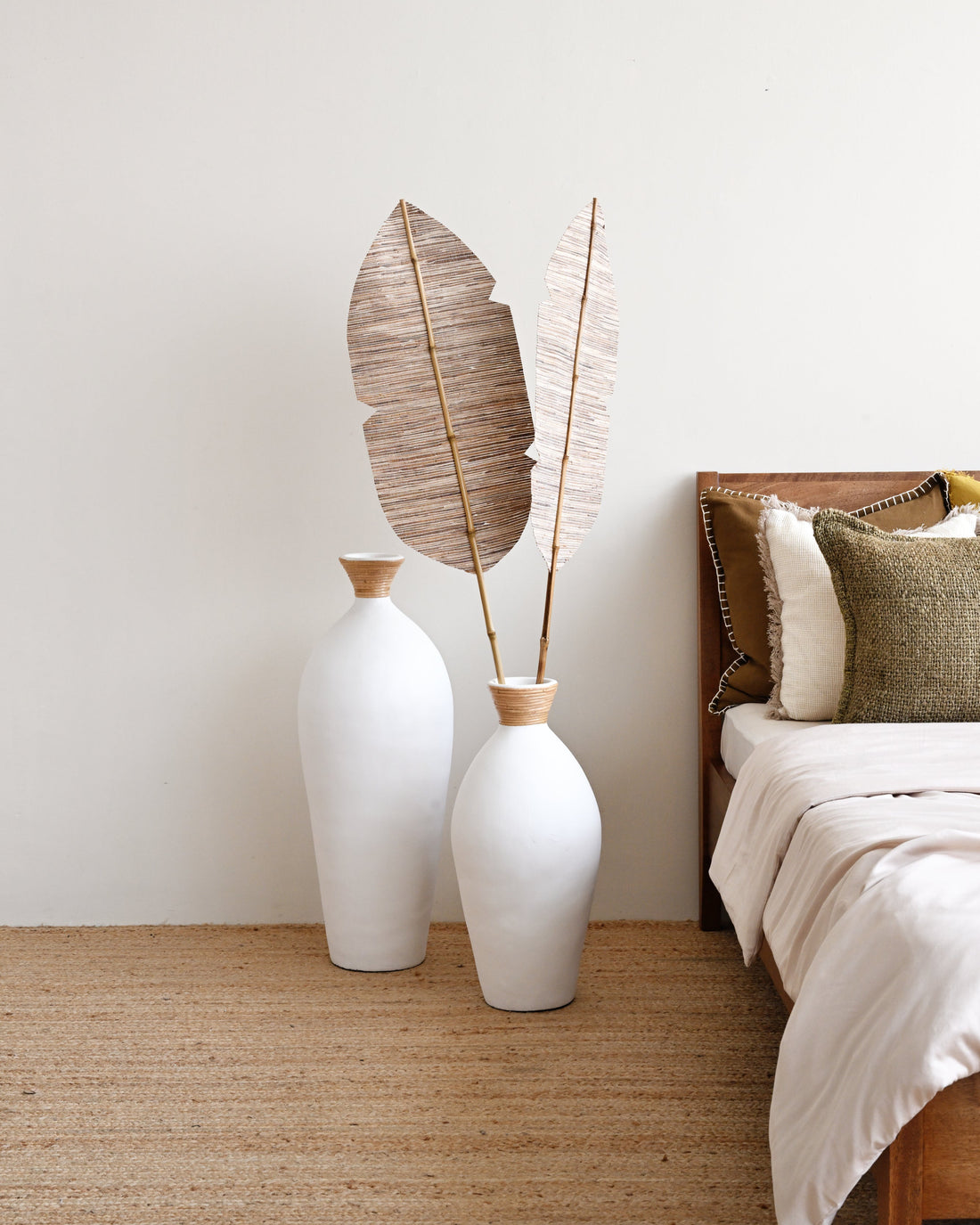 Two white vases with dried leaves on a wooden floor next to a bed with pillows.
