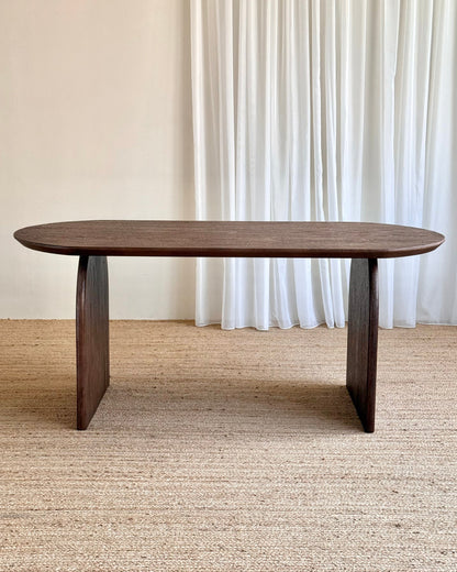 A dark brown wooden dining table with a rounded rectangular top and blocky pedestal legs, set on a textured beige jute rug in front of white curtains.