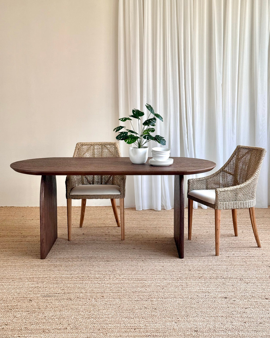 A dark brown oval wooden dining table with a thick block base, styled with two woven wicker chairs and a small potted plant on a neutral beige rug in front of white curtains.