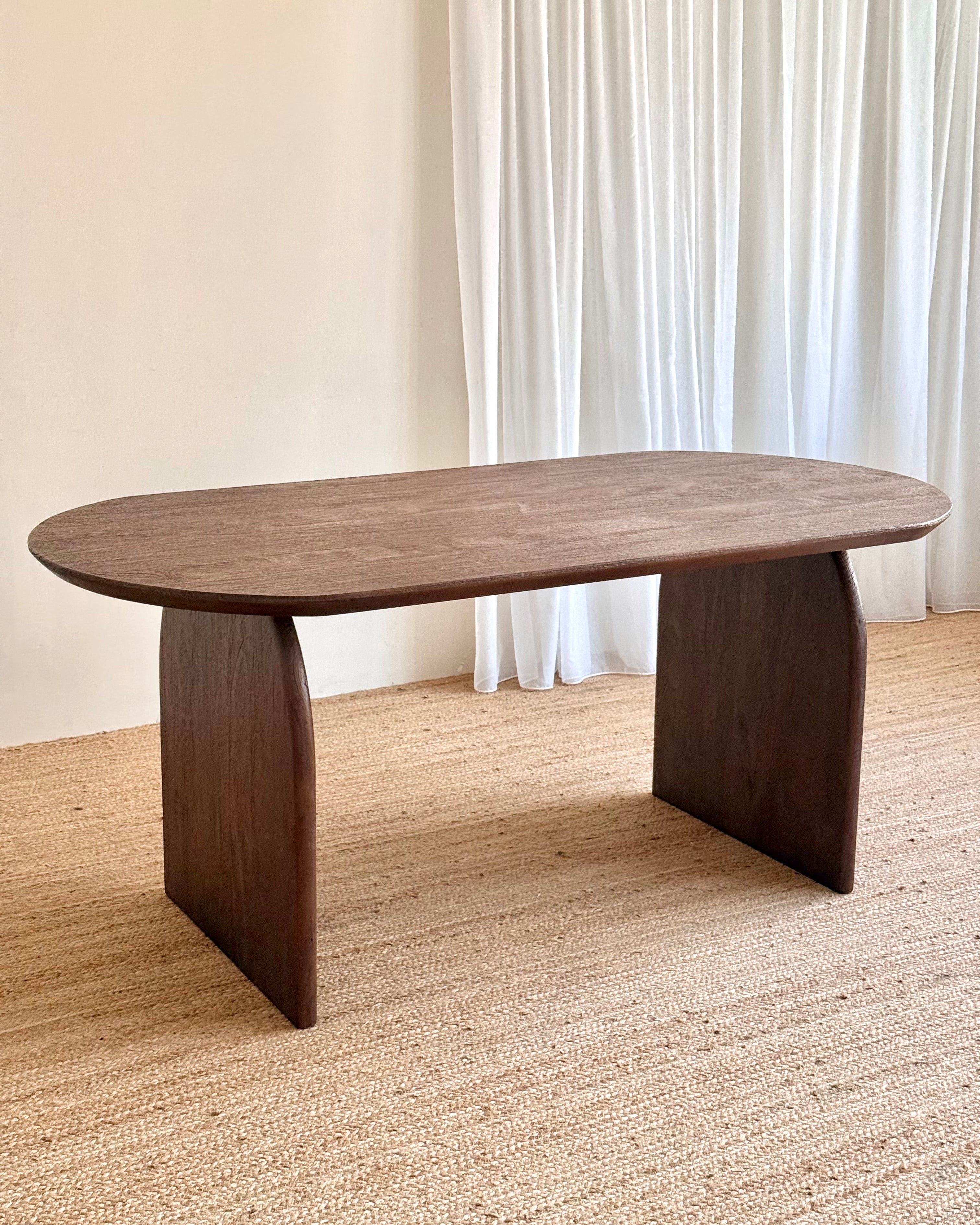 A dark brown wooden dining table with a rounded rectangular top and two integrated pedestal legs, set on a woven jute rug in front of white curtains.