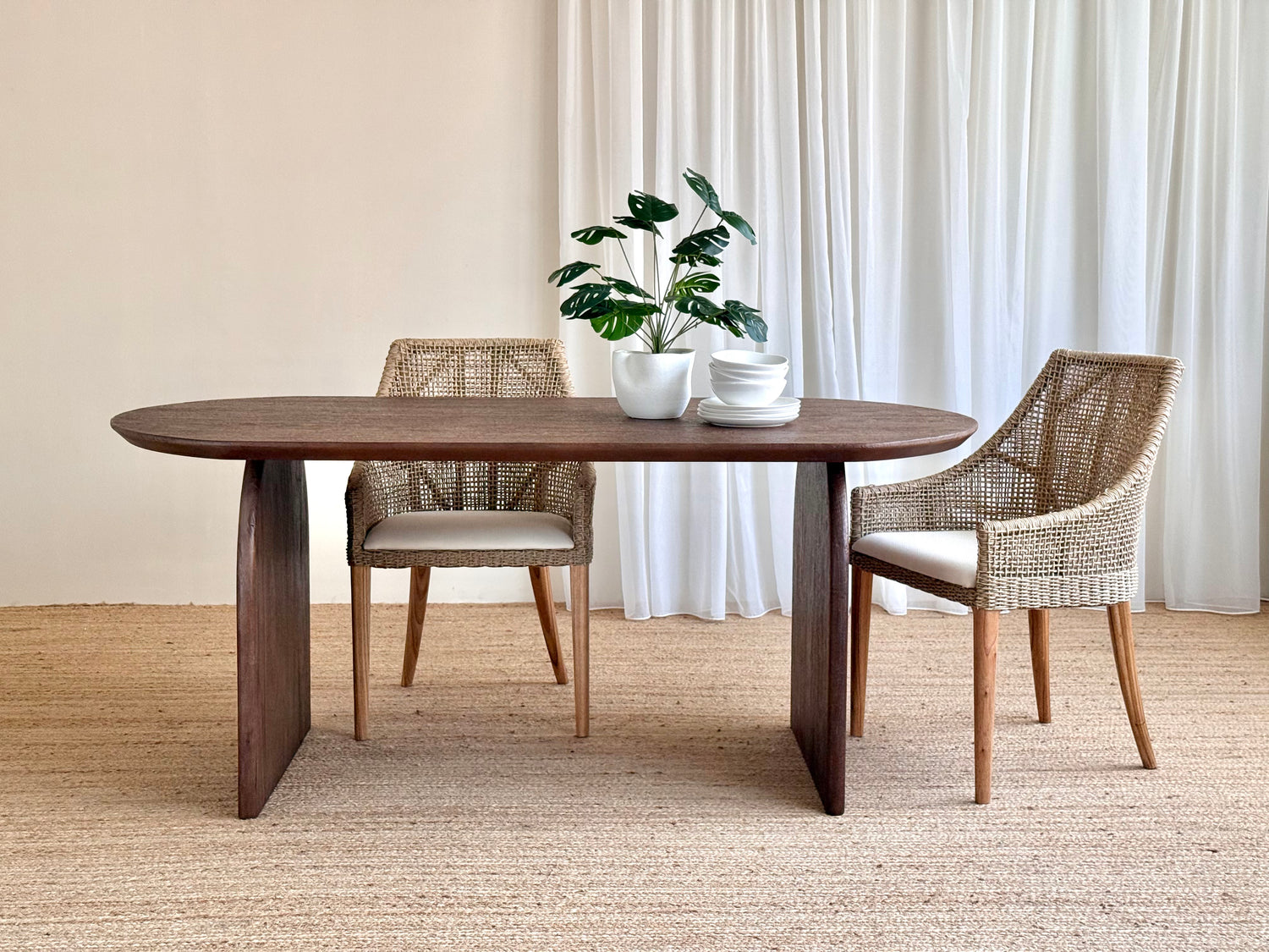 A dark brown oval wooden dining table with two woven wicker chairs, styled with a potted plant and white bowls on a neutral beige background.