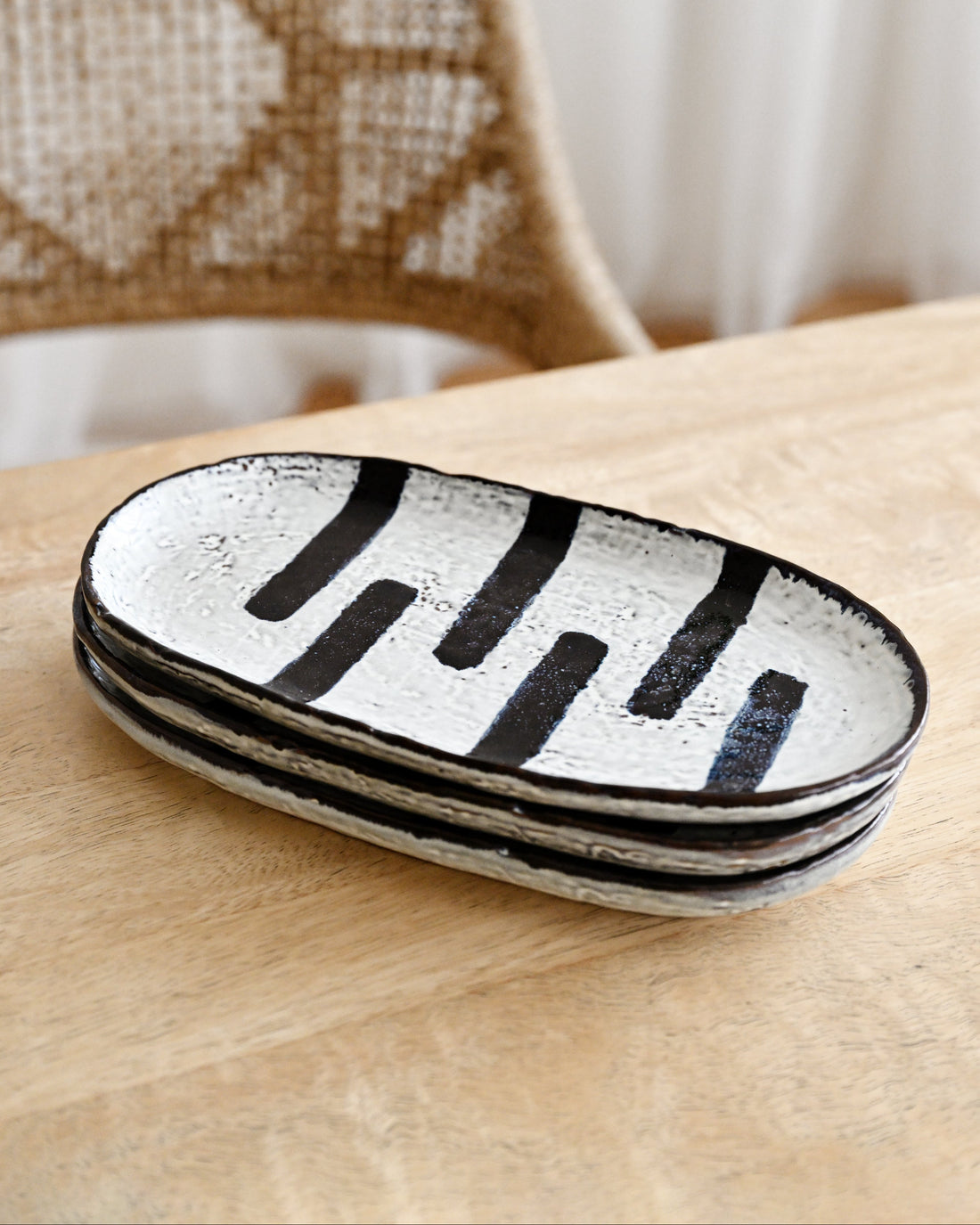 Stack of black and white ceramic trays on a wooden surface with a wicker chair in the background.