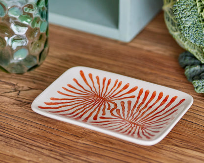 Decorative plate with red pattern on a wooden surface, glass, and plant in the background