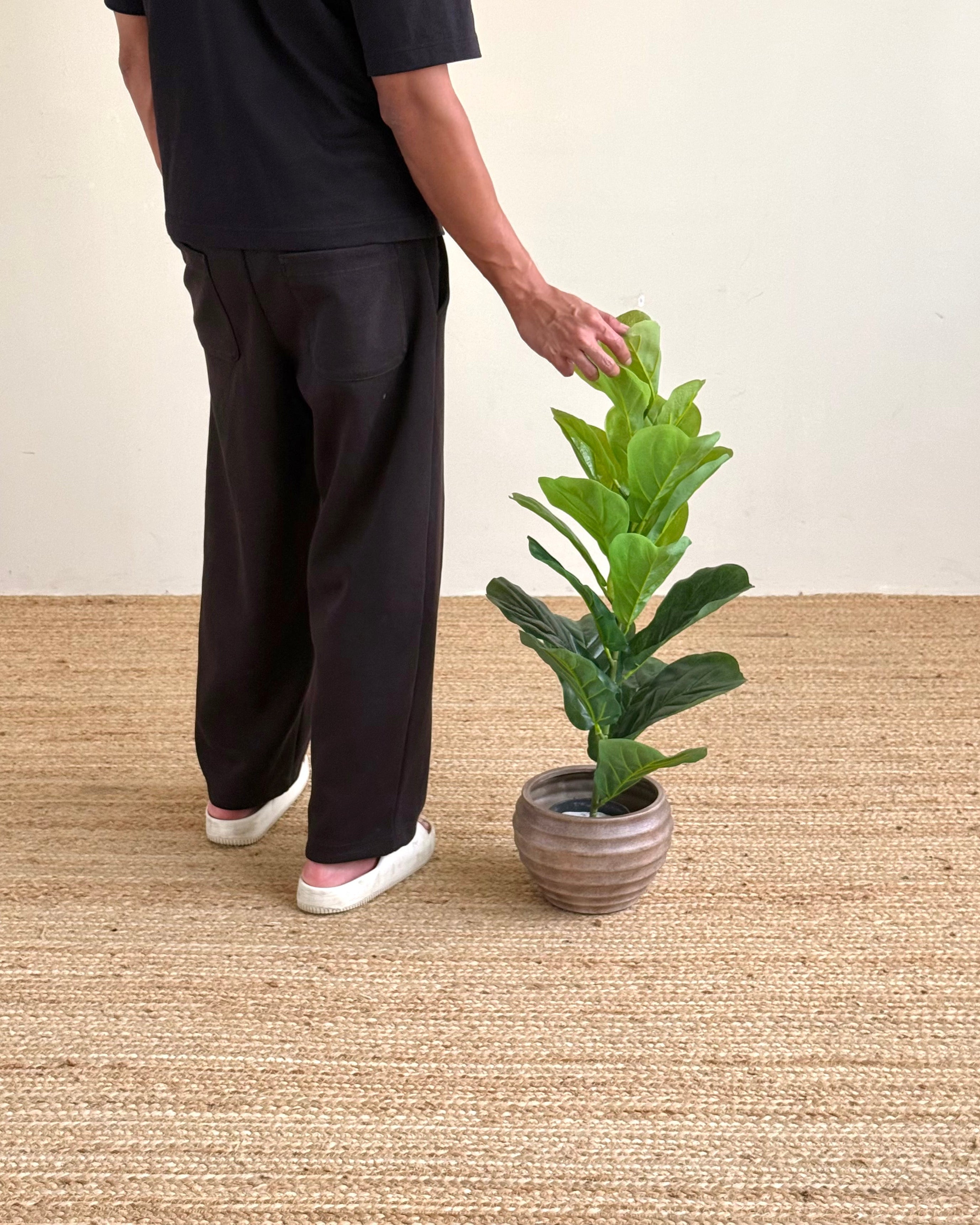 A person wearing a black t-shirt and black wide-leg pants stands beside a potted green plant on a woven beige rug against a plain cream wall.