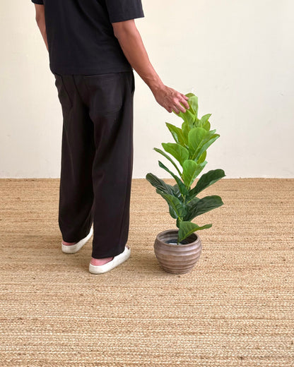 A person wearing a black t-shirt and black wide-leg pants stands beside a potted green plant on a woven beige rug against a plain cream wall.
