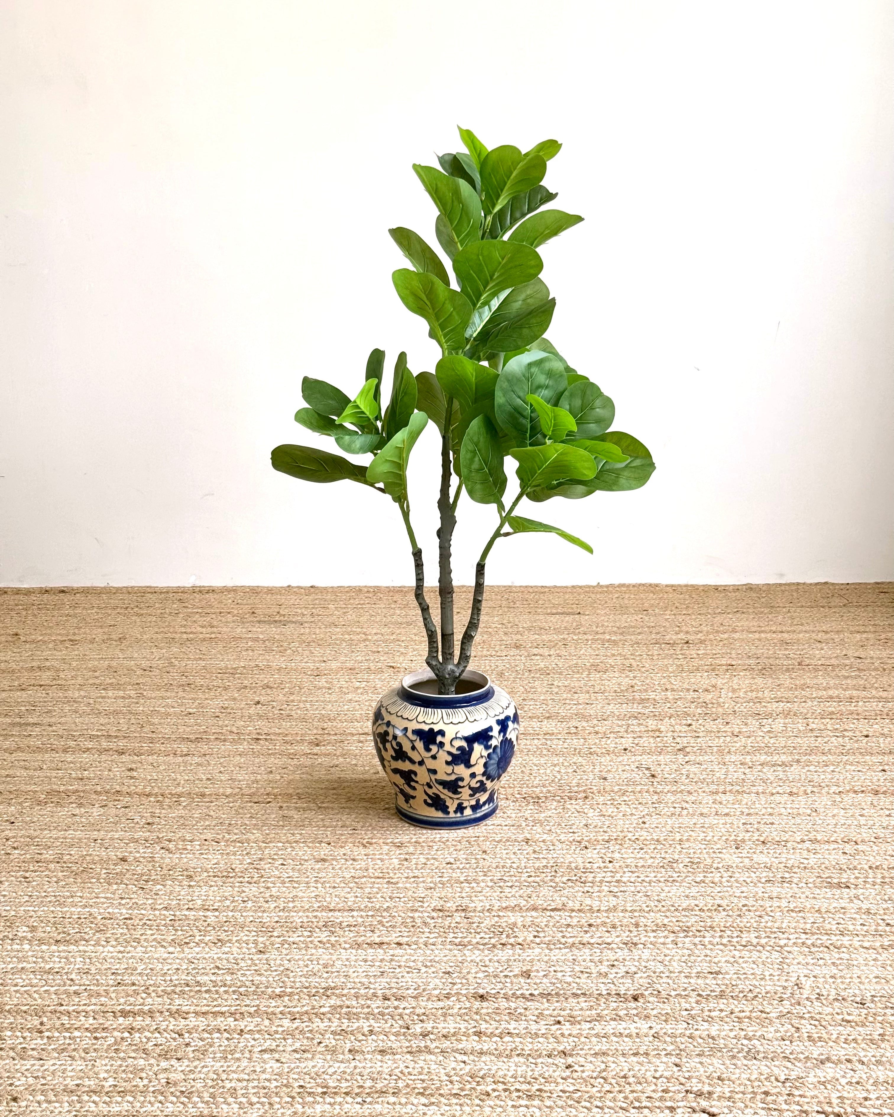 An artificial fiddle-leaf fig plant with green leaves and brown stems, potted in a blue and white ceramic jar, set on a beige woven rug against a plain white wall.