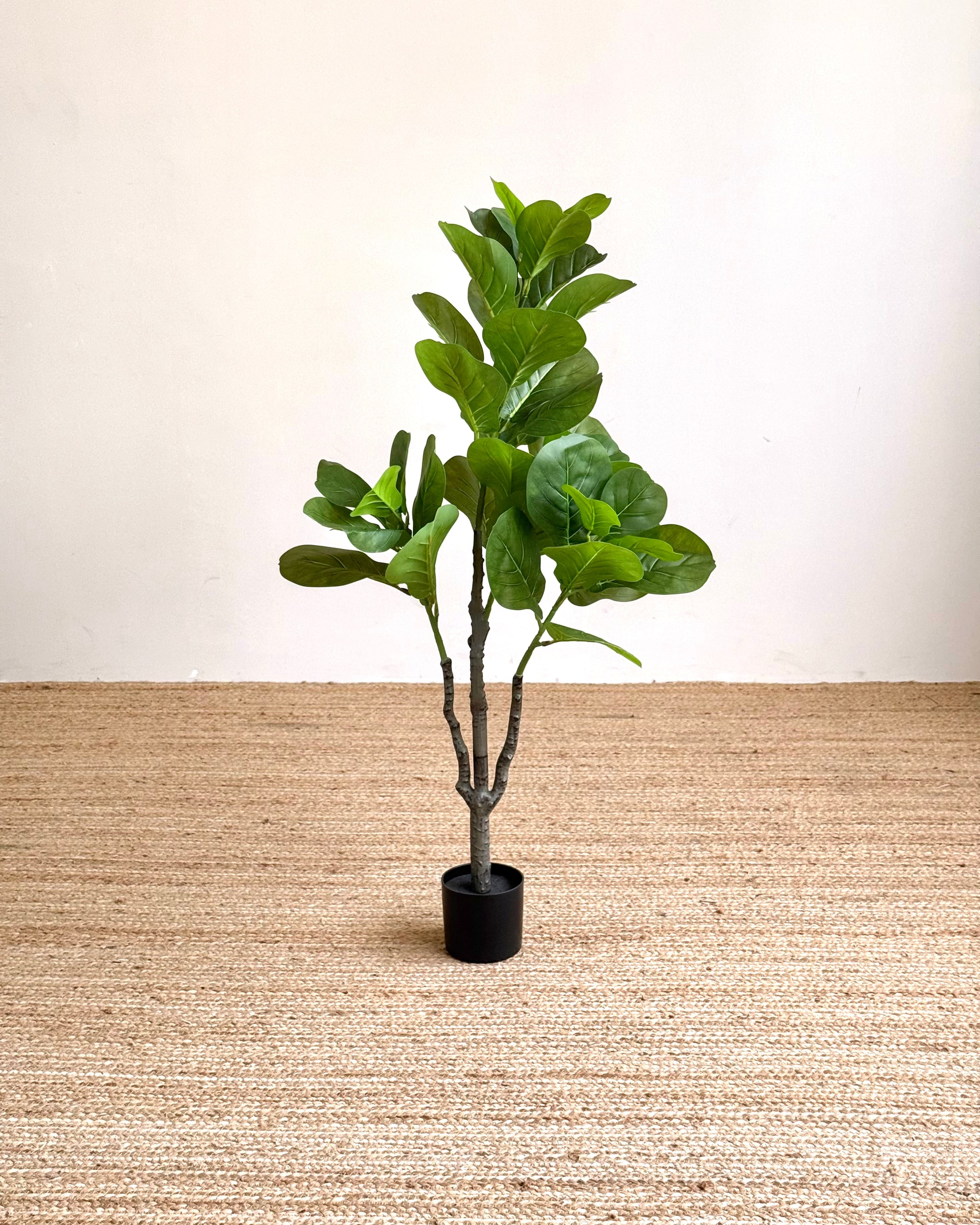 An artificial fiddle-leaf fig plant with green leaves in a black pot, placed on a woven jute rug against a plain off-white wall.