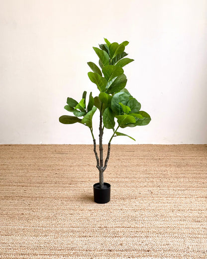 An artificial fiddle-leaf fig plant with green leaves in a black pot, placed on a woven jute rug against a plain off-white wall.