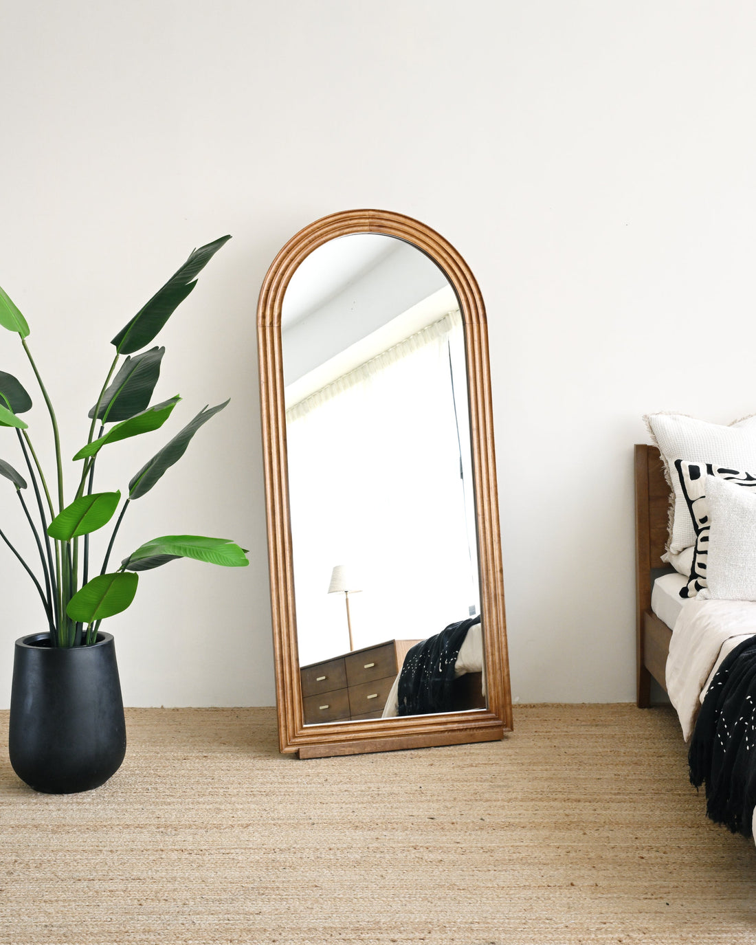 Wooden floor mirror leaning against a white wall in a bedroom with a plant and bed.