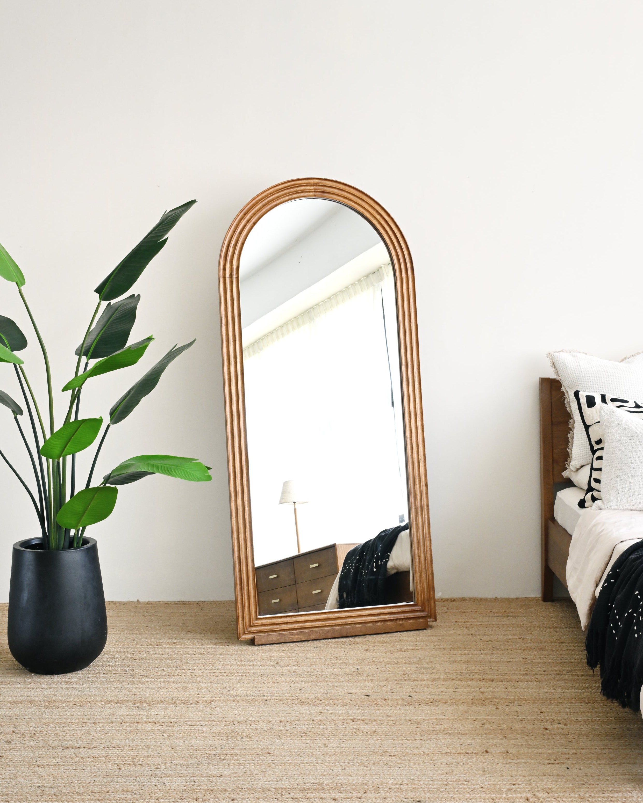 Wooden floor mirror leaning against a white wall in a bedroom with a plant and bed.